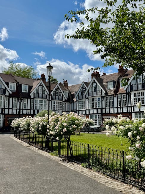 A picturesque residential garden in a terraced neighborhood during daytime, featuring a well-maintained lush green lawn bordered by a black wrought iron fence, with blooming white rose bushes lining t